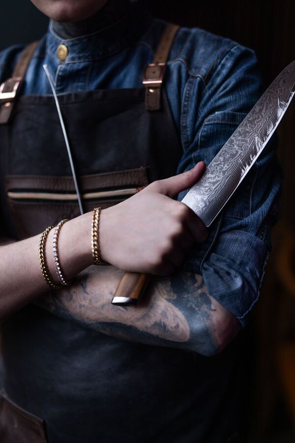 Chef with tattooed arms holding a large kitchen knife against a denim apron