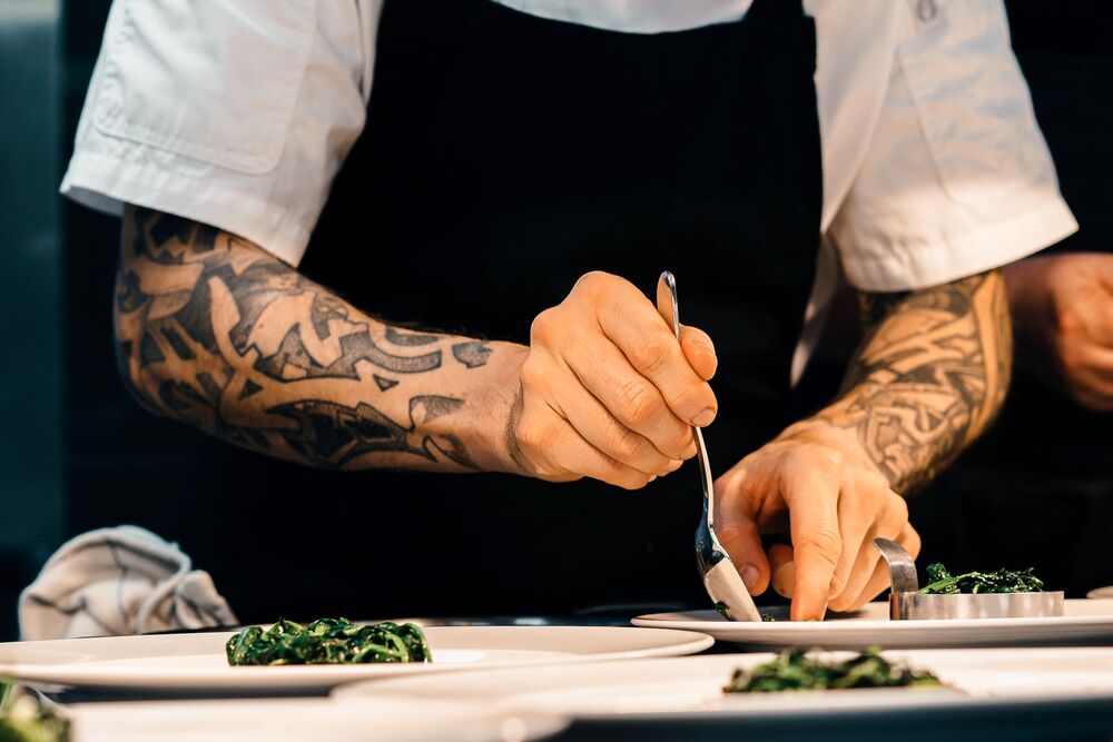 Chef with tattooed forearms plating a dish with tweezers