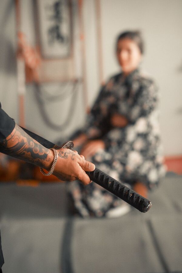 Tattooed hand holding a katana with a person in traditional dress behind