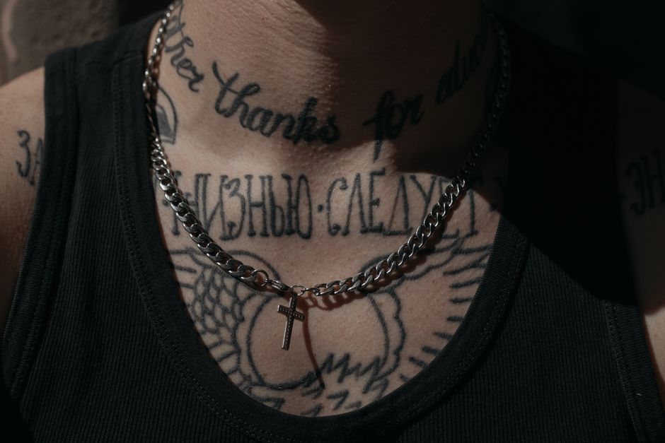 Script lettering tattoos across a person's collarbone and chest, with a silver cross pendant