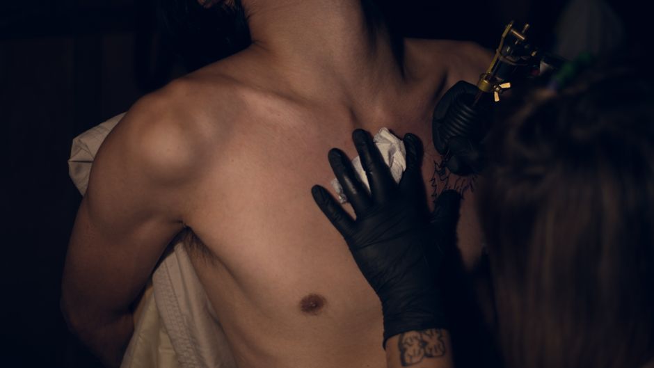 An artist tattoos a man's chest in a dimly lit tattoo shop setting.