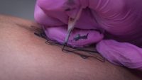 Close up of a tattoo needle inking black linework into skin, held in a gloved hand