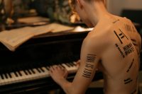 Person with bold black lettering on their back and arm playing a piano beside sheet music