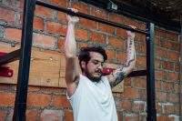 Hombre con los brazos tatuados haciendo dominadas en un gimnasio con pared de ladrillo