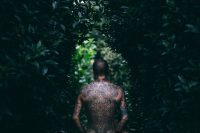 Man with an ornamental blackwork back piece walking between dark green trees