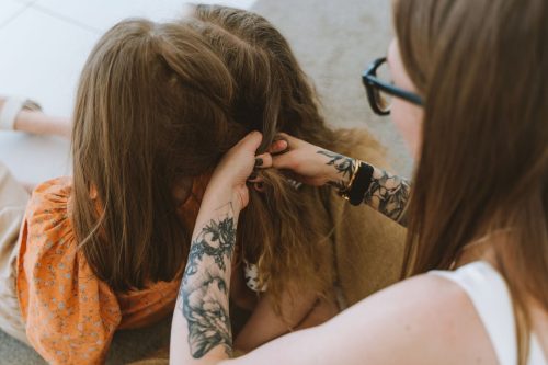 Madre con los brazos tatuados trenzando el pelo de su hija en casa