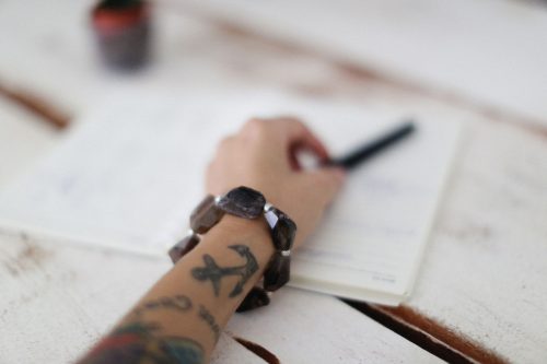 Brazo tatuado con pulsera escribiendo en un cuaderno abierto sobre una mesa de madera