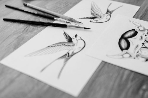 Bocetos en blanco y negro de golondrinas para tatuaje sobre una mesa de madera con lápices