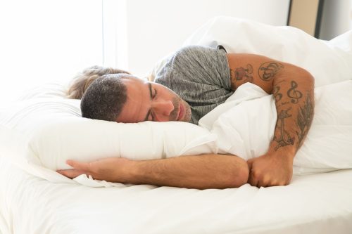 Hombre con el brazo tatuado durmiendo boca abajo sobre una almohada blanca