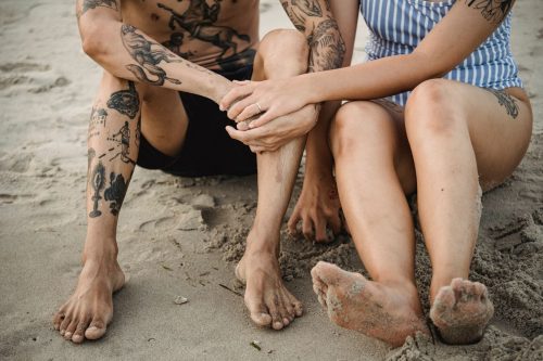 Pareja con tatuajes en brazos y piernas sentada descalza en la arena de la playa